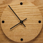 A square wall clock with round face, black arms and 4 black dots to indicate the time.