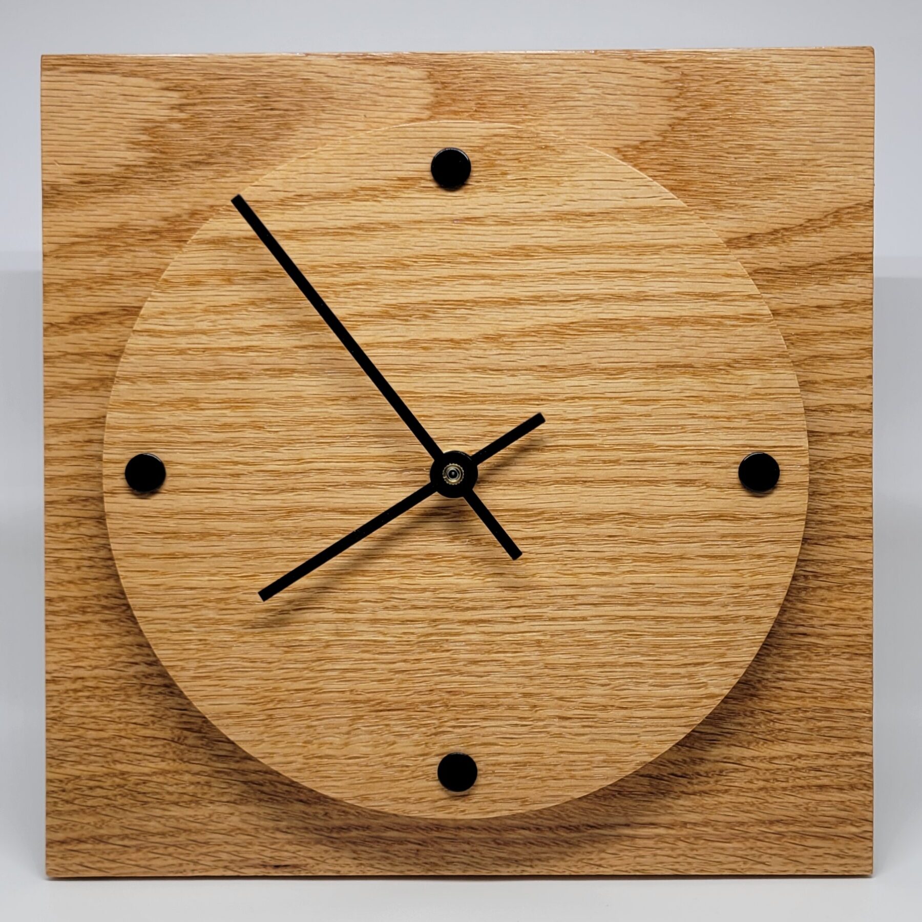 A square wall clock with round face, black arms and 4 black dots to indicate the time.