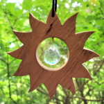 A wood suncatcher hanging in a window. Shaped like the sun, a glass prism ball is suspended in the middle.