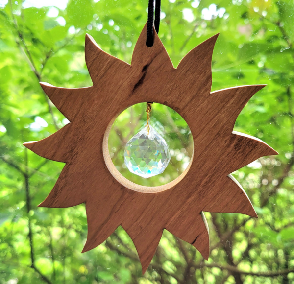 A wood suncatcher hanging in a window. Shaped like the sun, a glass prism ball is suspended in the middle.