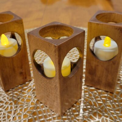 Three simple candle holders made from rectangular blocks of wood, with a hole on each of the four sides and the top. Flameless candles sit inside the blocks