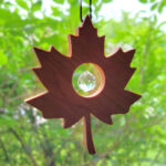 A solid wood, maple leaf shaped suncatcher, hangs in a window with green trees behind. It has a glass prism ball nestled in the middle of the leaf.