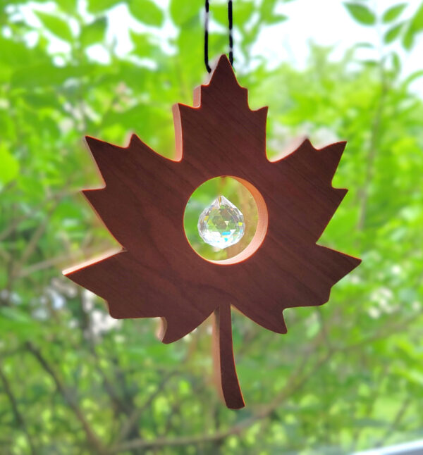 A solid wood, maple leaf shaped suncatcher, hangs in a window with green trees behind. It has a glass prism ball nestled in the middle of the leaf.