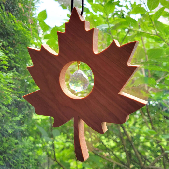 A solid wood, maple leaf shaped suncatcher, hangs in a window with green trees behind. It has a glass prism ball nestled in the middle of the leaf.