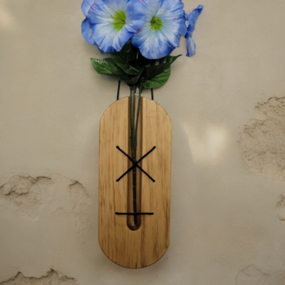 A glass, test tube, nestled in a solid piece of pine, held in place by brown lacing. This hangs on a wall with a blue flower extending from the top of the tube.