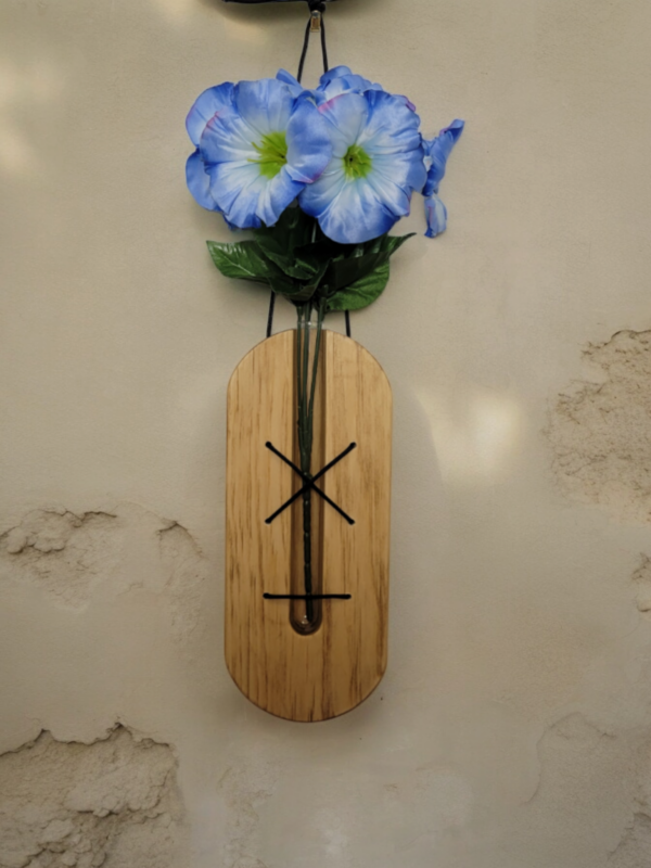 A glass, test tube, nestled in a solid piece of pine, held in place by brown lacing. This hangs on a wall with a blue flower extending from the top of the tube.