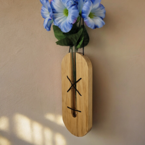 A glass, test tube, nestled in a solid piece of pine, held in place by brown lacing. This hangs on a wall with a blue flower extending from the top of the tube.