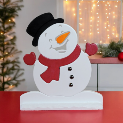 A wooden snowman ornament sitting on a table. He is white with a black hat, red scarf and mits, black buttons and a carrot nose.