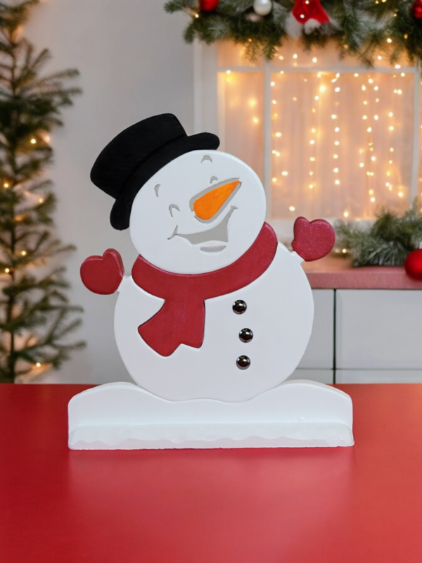 A wooden snowman ornament sitting on a table. He is white with a black hat, red scarf and mits, black buttons and a carrot nose.