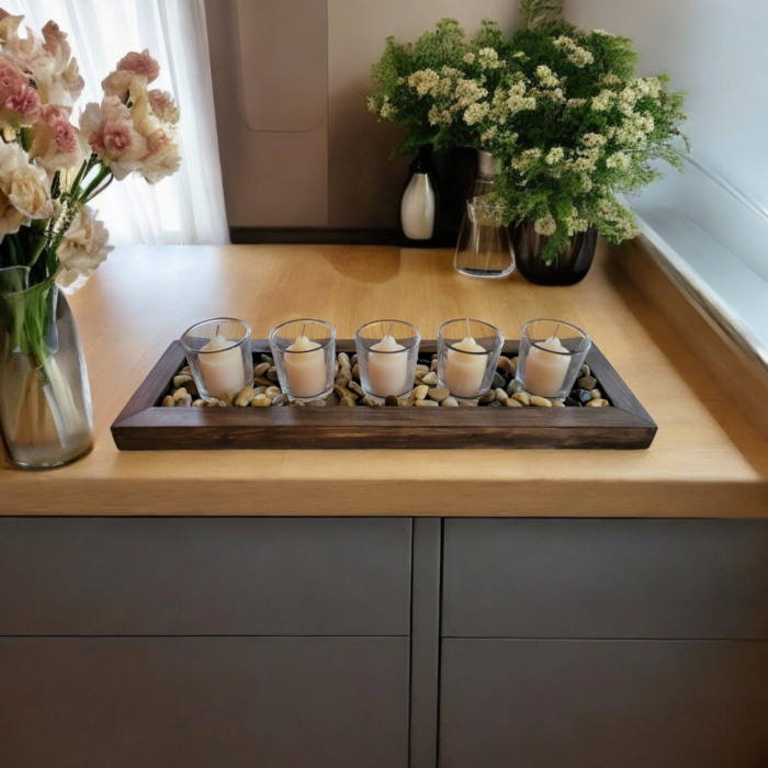 A simple rectangular wooden tray, covered with small smooth stones. Nestled among the stones are five glass votive candle holders.