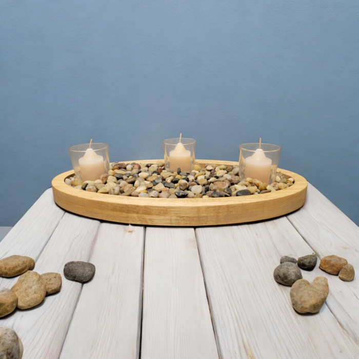 A simple oval wooden tray, covered with small smooth stones. Nestled among the stones are three glass votive candle holders.