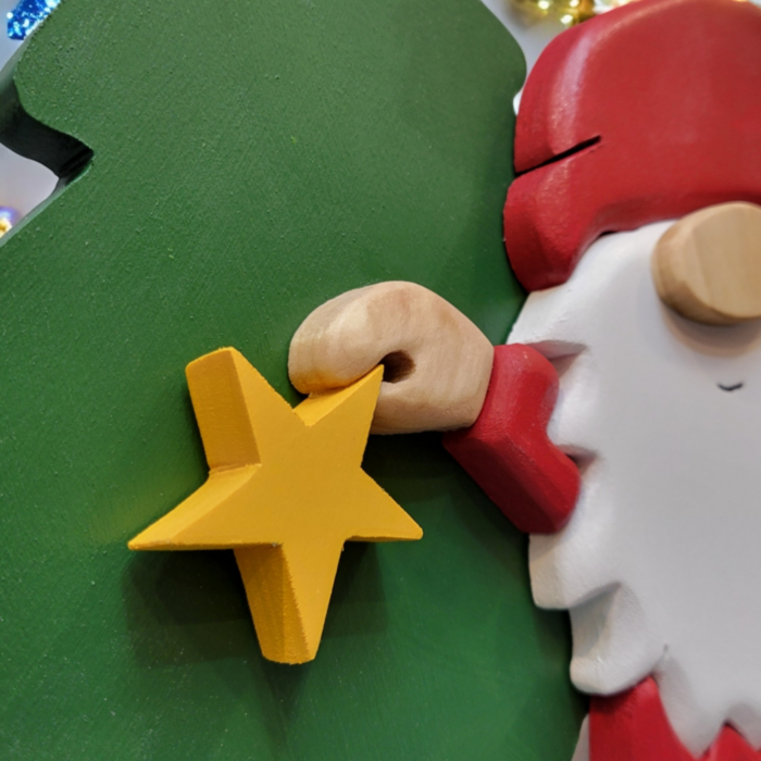 Christmas gnome with while beard, red hat, holding a yellow star, standing in front of a tall green tree.