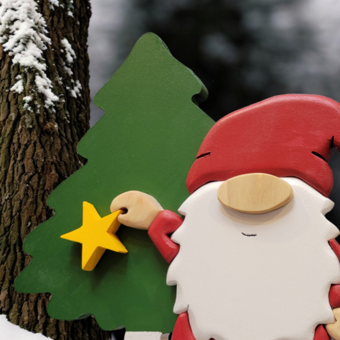 Christmas gnome with while beard, red hat, holding a yellow star, standing in front of a tall green tree.