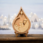 A christmas scene of ornaments and lights, with a wood teardrop shaped centerpiece depicting the Holy Family under the Star.