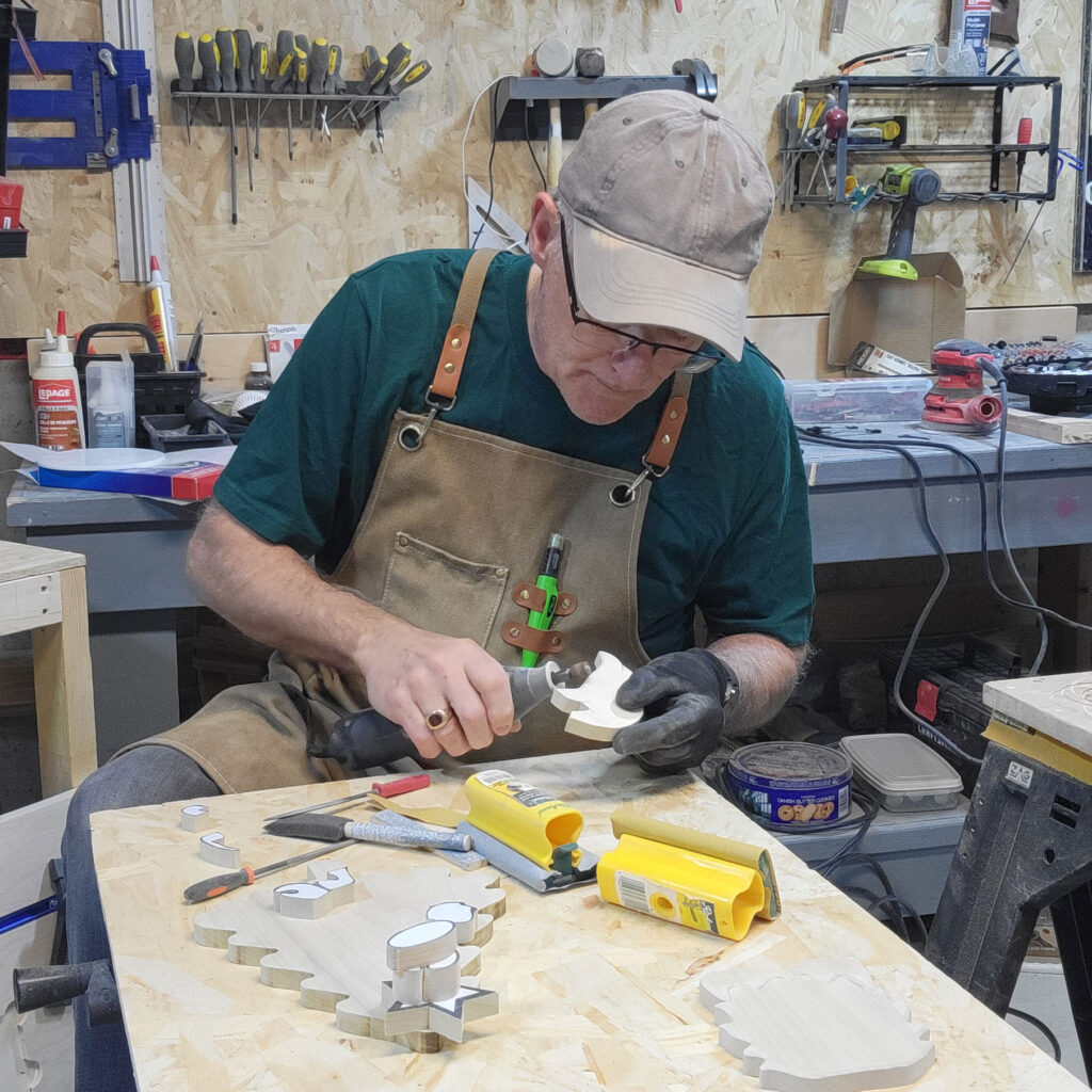 Shop owner, Michael, sitting at the workbench, shaping a piece of wood, using a drill-like sanding tool.